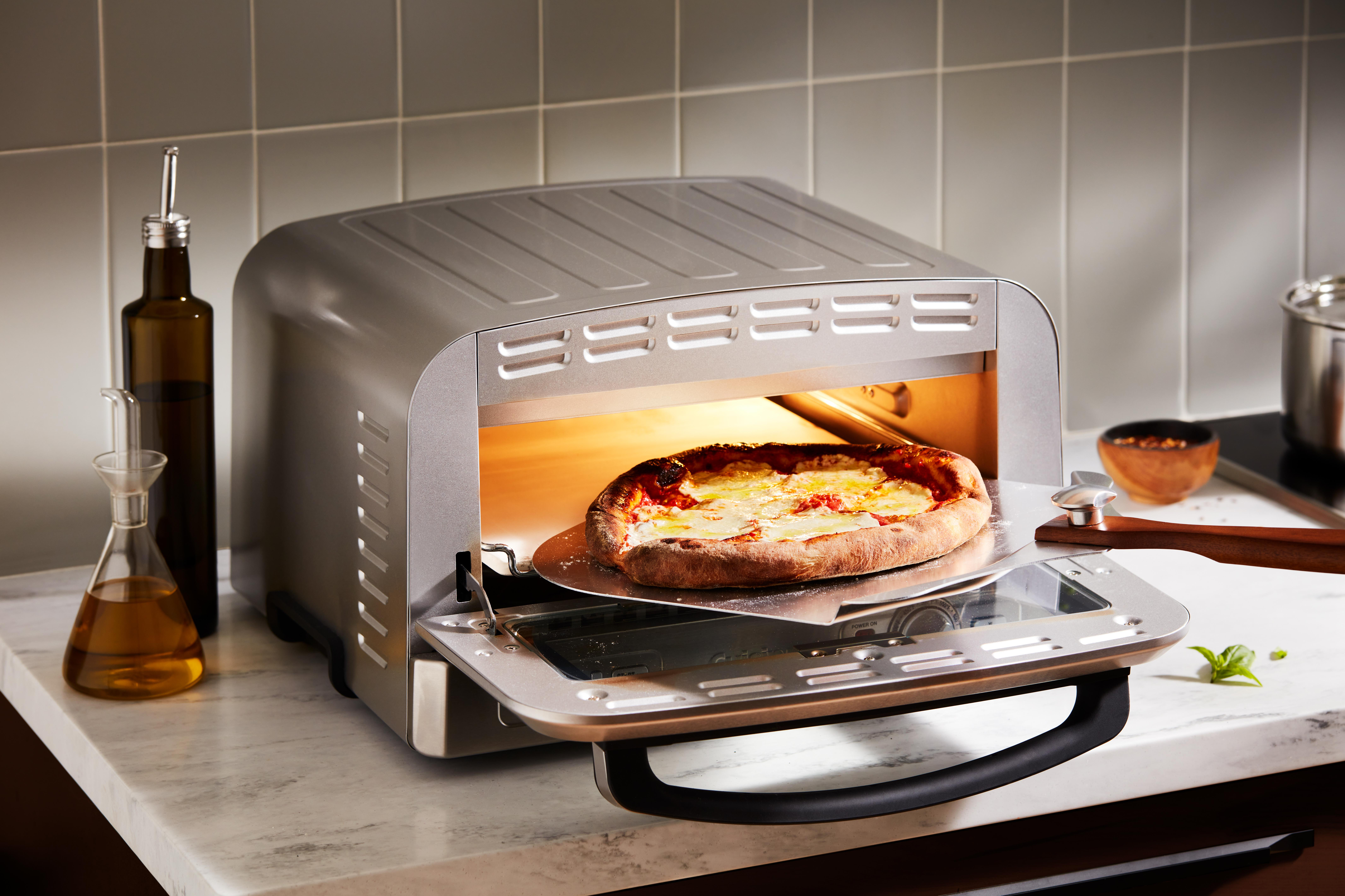 Eine Pizza wird in einem silbernen Toaster gebacken. Olivenölflaschen und eine kleine Schüssel stehen auf der Arbeitsplatte.