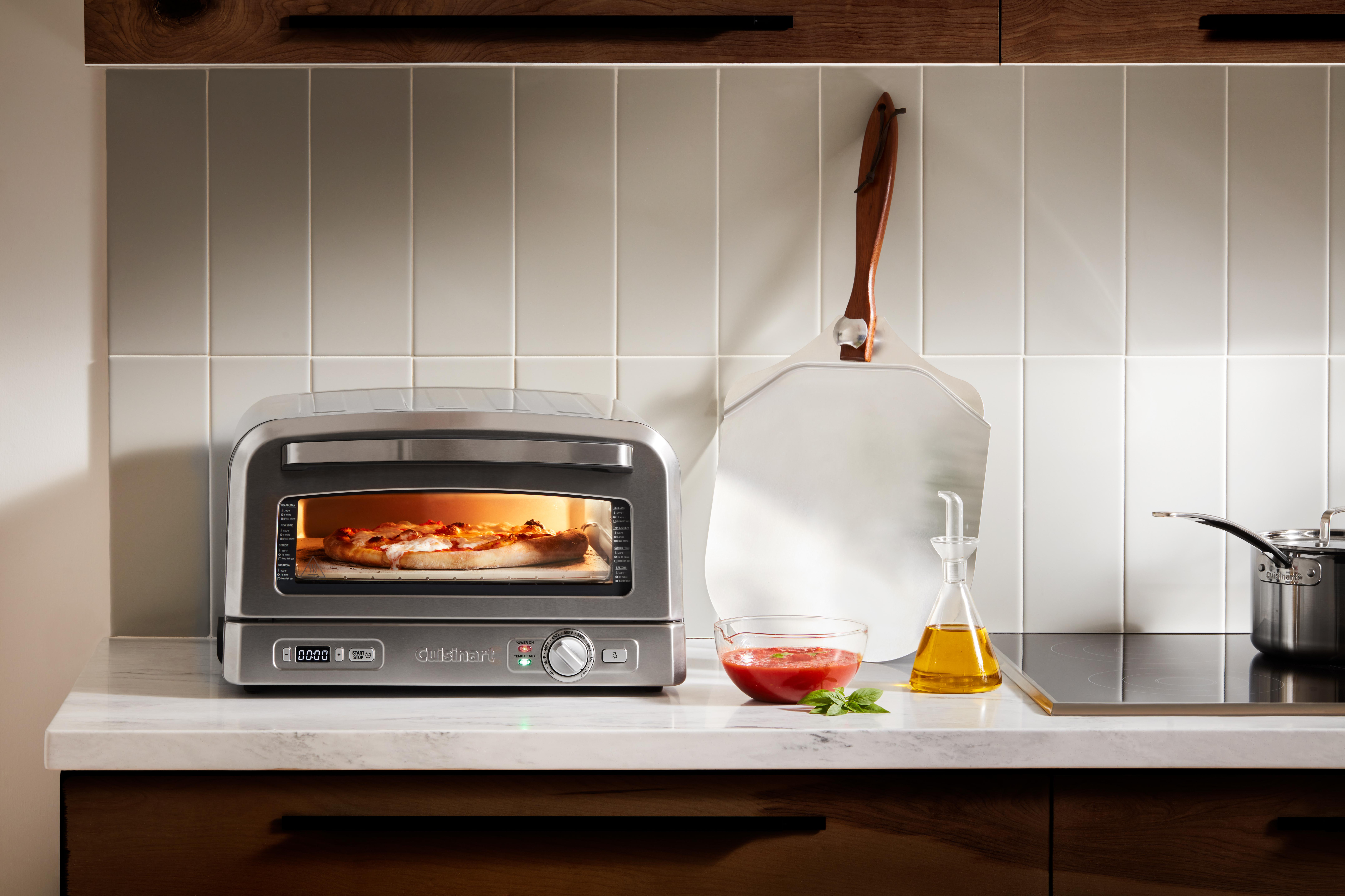 Ein Pizzaofen mit Pizza, daneben Zutaten und Küchenutensilien. Der Hintergrund ist eine weiße Kachelwand.