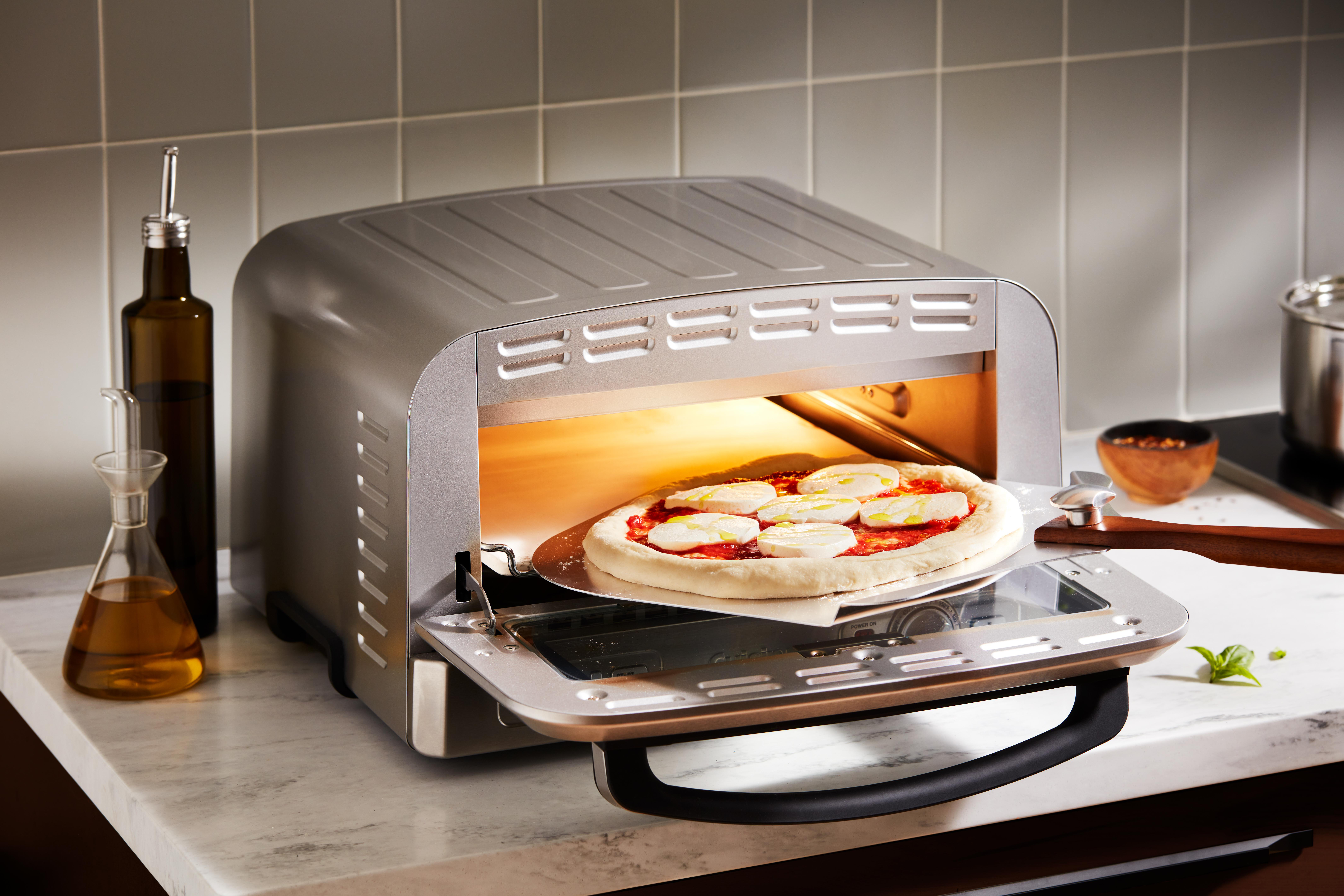 Eine Pizza, die in einem silbernen Ofen gebacken wird. Zutaten sind Mozzarella, Tomatensauce und Olivenöl. Andere Gegenstände auf der Theke.