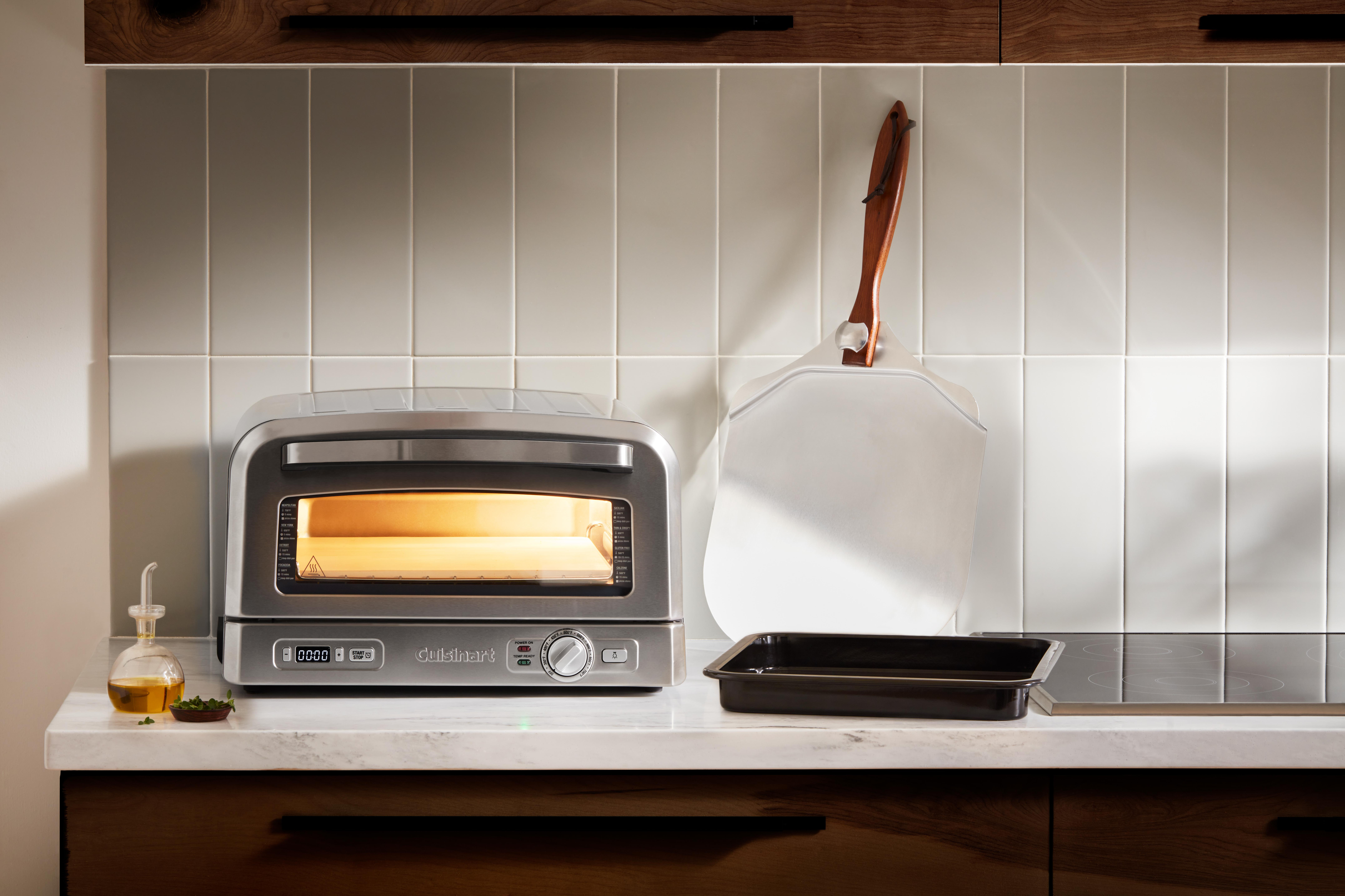 Ein Edelstahl-Toaster, Pizzaschieber und Backblech stehen auf einer Theke. Ölflasche und Kräuter daneben.