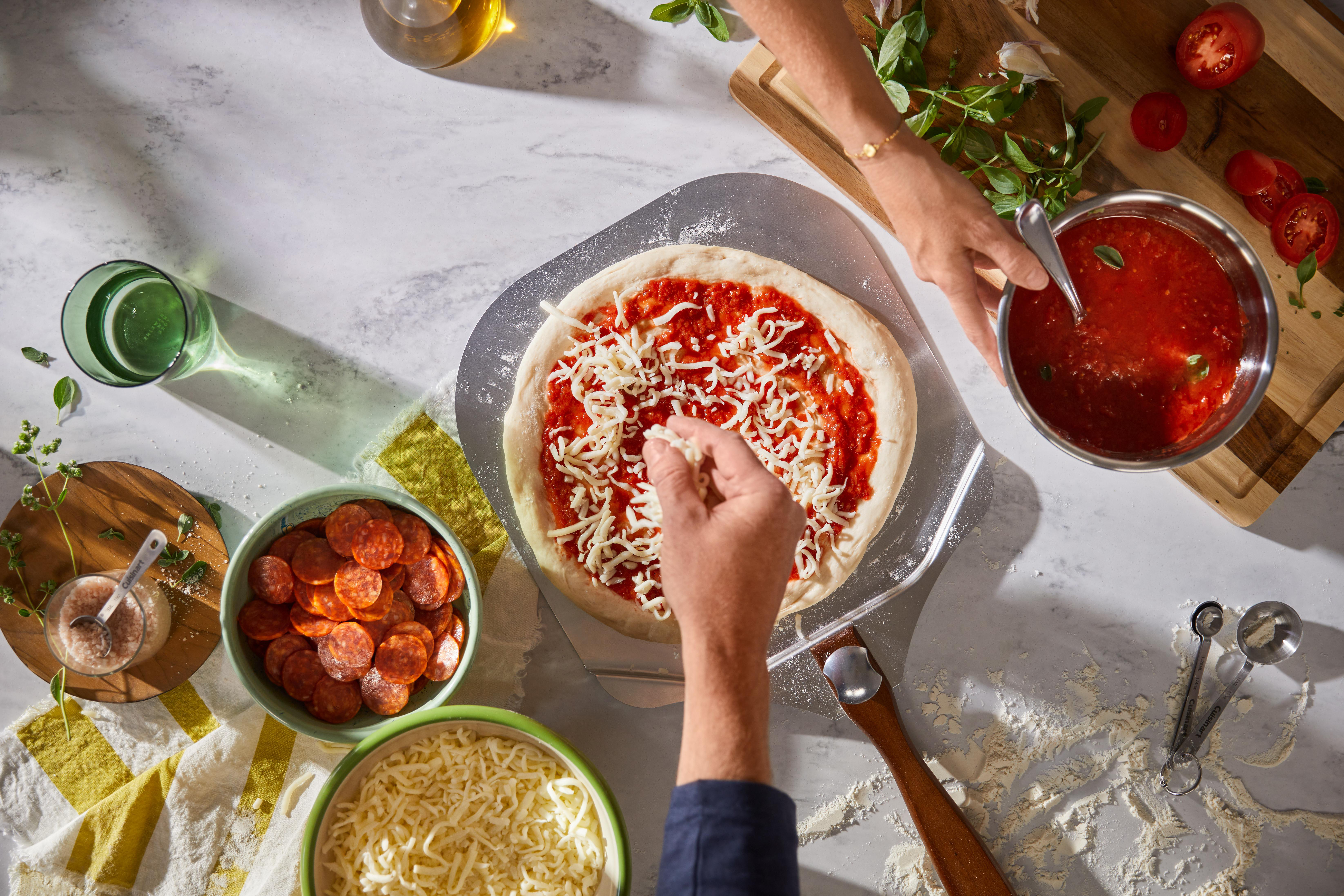 Hände bereiten Pizza zu. Pizzateig mit Soße und Käse. Zutaten wie Peperoni, Käse, Tomaten und Kräuter auf einer Marmoroberfläche.