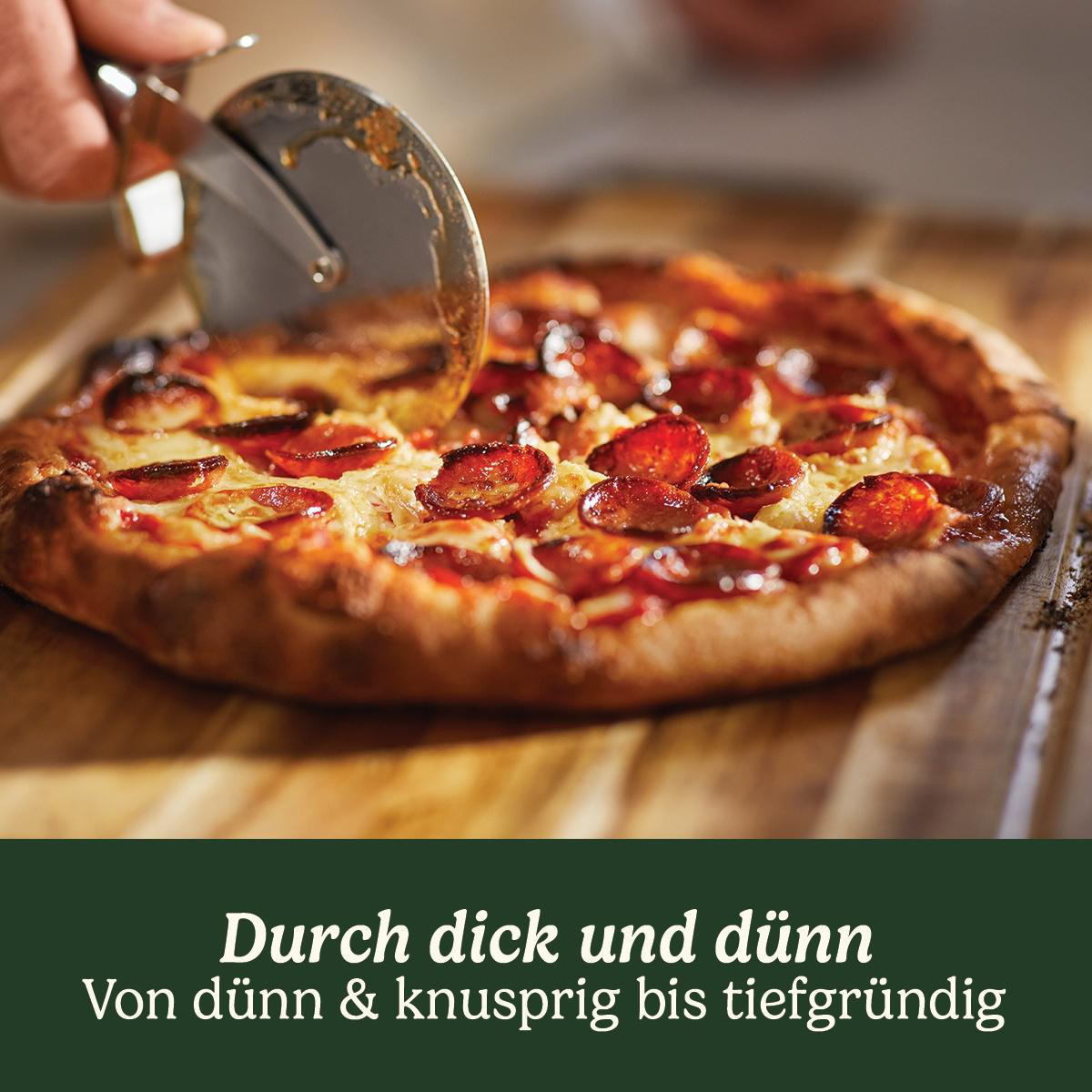 Eine Pizza wird mit einem Pizzaschneider geschnitten. Sie hat Pepperoni und Käse. Der Teig ist goldbraun, auf einem Holzbrett.