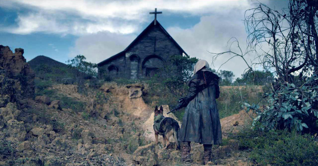 Eine Person und ein Hund stehen vor einer Kirche mit Kreuz. Die Szene hat eine unheilvolle Atmosphäre mit einem bewölkten Himmel.