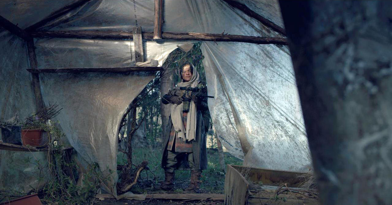 Eine Person steht in einem provisorischen Unterstand und hält ein Gewehr. Der Unterstand besteht aus Holz und Plastikfolie, in einem Wald.