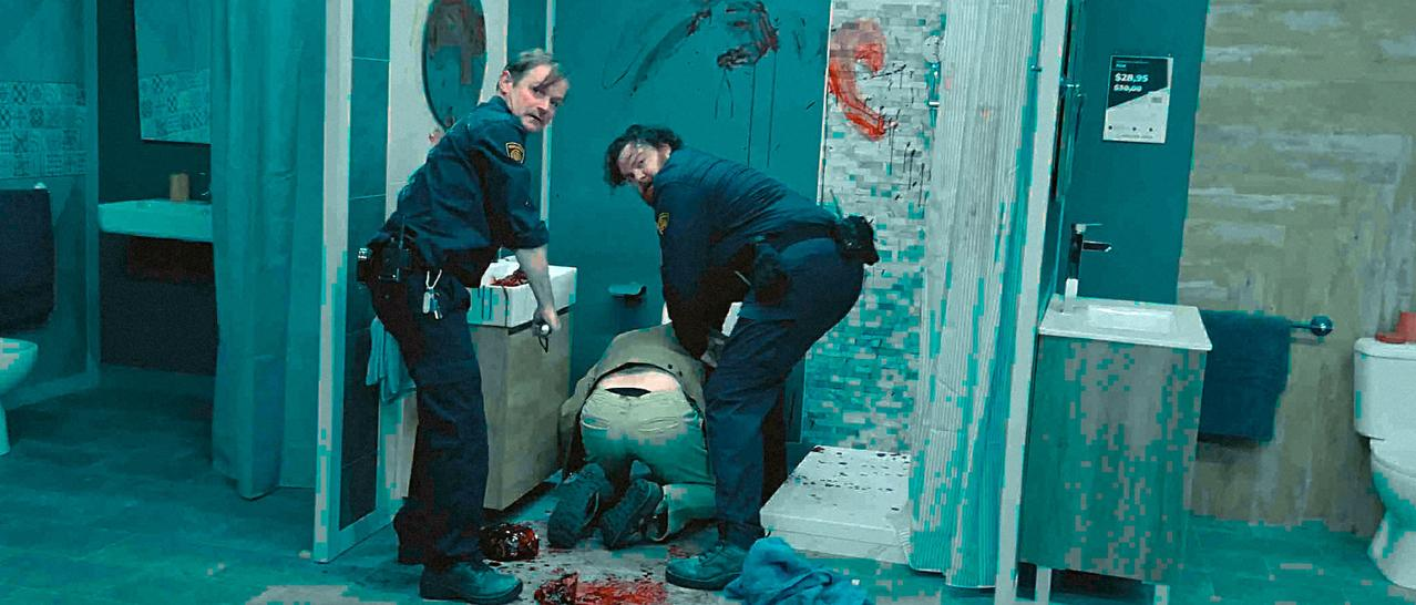 Zwei Polizisten untersuchen eine Person auf dem Badezimmerboden. Blut ist auf dem Boden und an der Wand verspritzt. Die Szene ist düster.