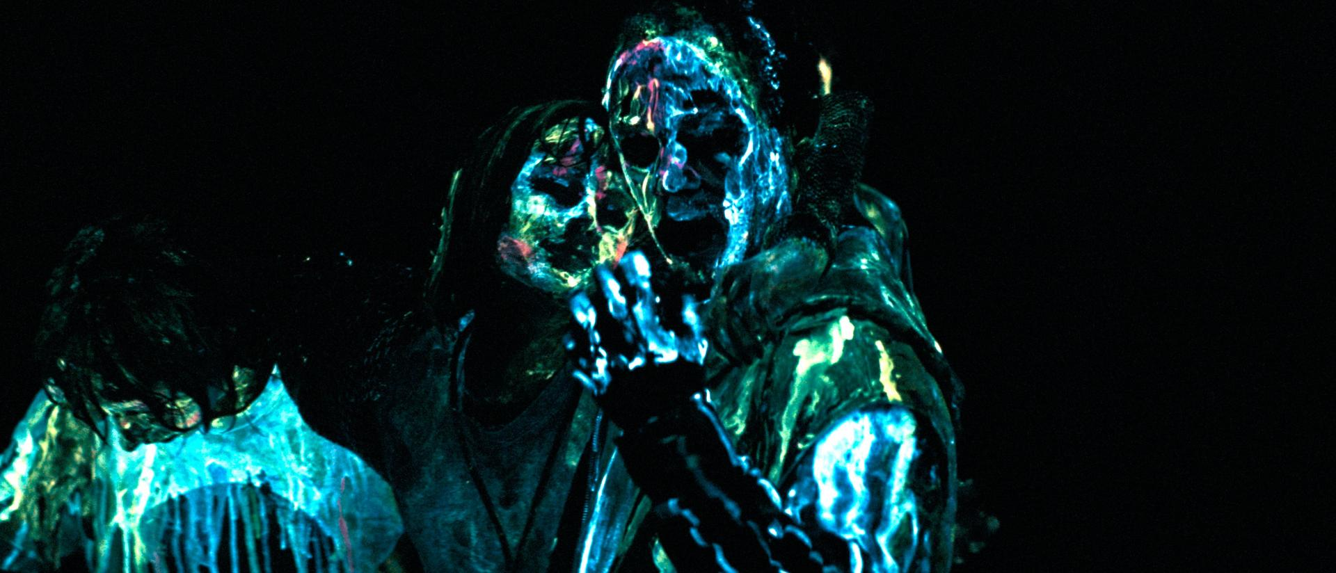 Drei Figuren in dunkler Umgebung, beleuchtet mit blauem und grünem Licht. Gesichter sind sichtbar, mit offenem Mund. Die Szene hat einen mysteriösen Ton.