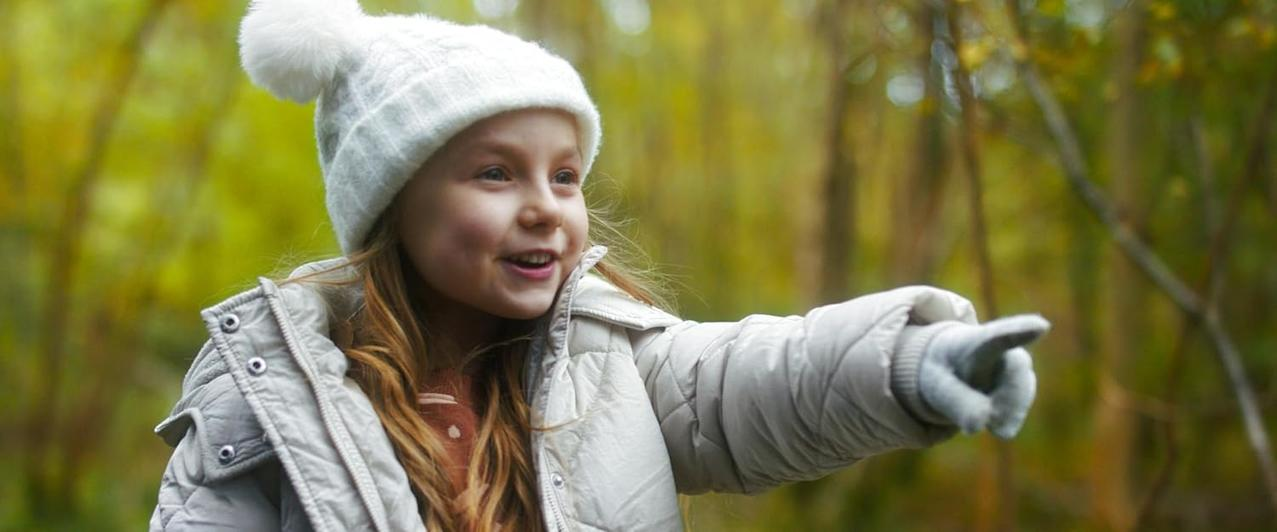 Ein Mädchen mit weißer Mütze und Jacke zeigt nach rechts und lächelt in einer herbstlichen Waldkulisse. Der Hintergrund ist unscharf.