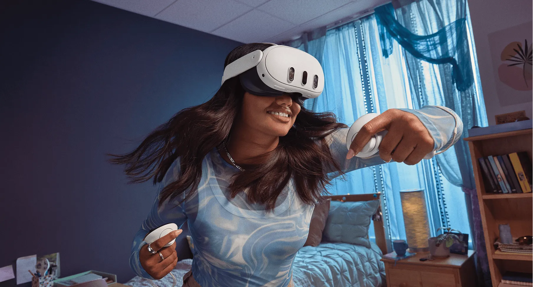 Donna con visore VR e controller, sorridente. Camera da letto con tende blu e letto sullo sfondo.