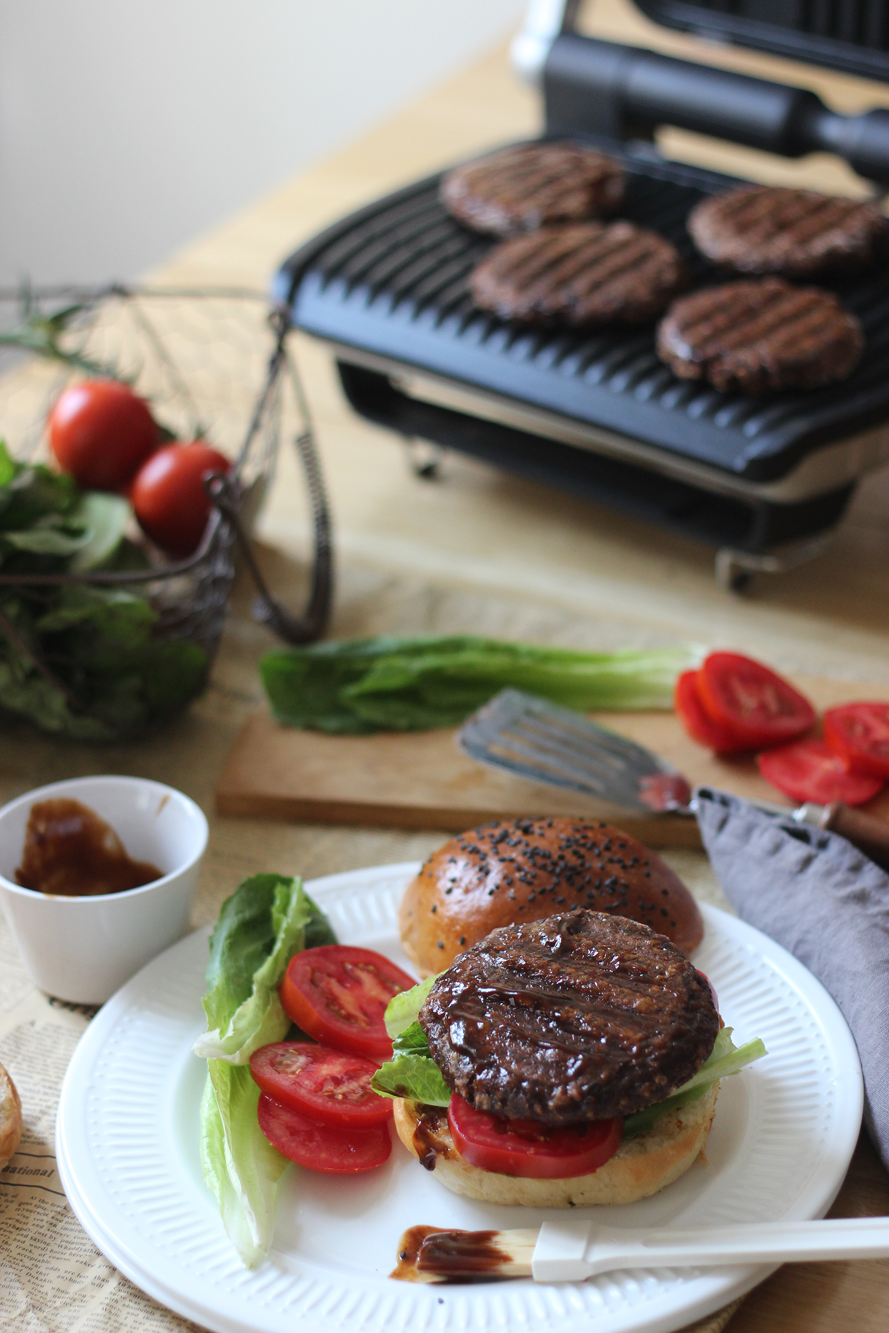 Hamburger paradicsommal és salátával, egy grill mellett, amin hamburgerek vannak. Paradicsom és zöldségek közelben.
