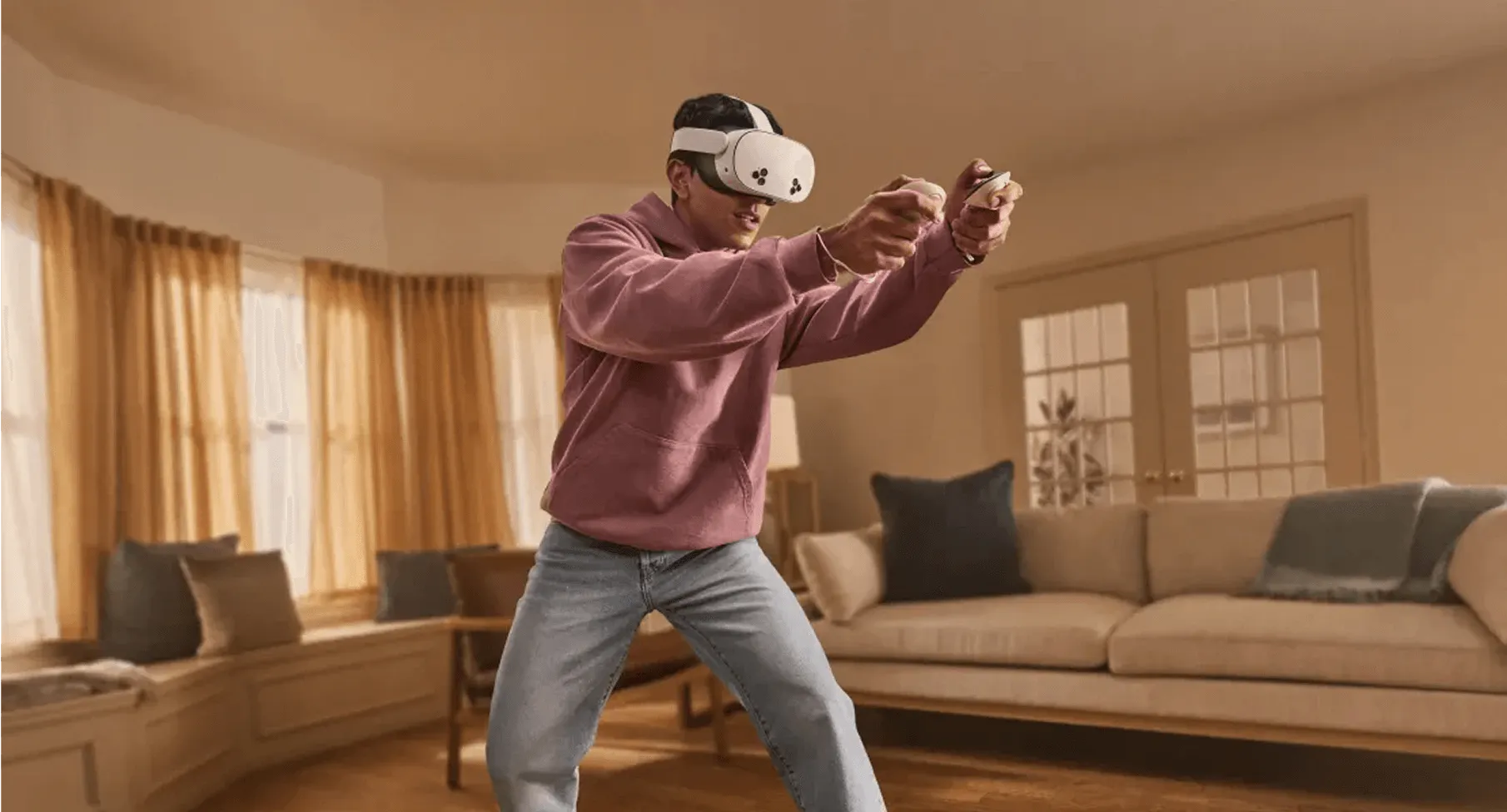 Un uomo con un visore VR e controller in un salotto. Indossa una felpa con cappuccio rosa e jeans.