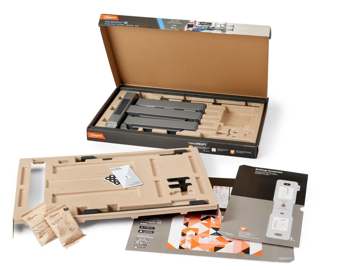 Otwarty karton z częściami uchwytu TV. Zawiera uchwyt, szablon, instrukcję i małe woreczki. Opakowanie kartonowe na białym tle.