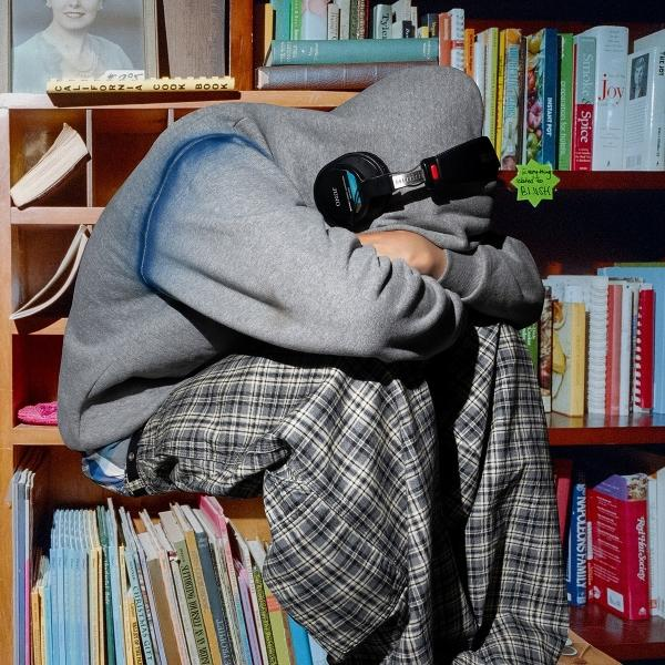 Eine Person in einem grauen Kapuzenpullover und einer karierten Hose sitzt vor einem Bücherregal und trägt Kopfhörer. Bücher sind sichtbar.
