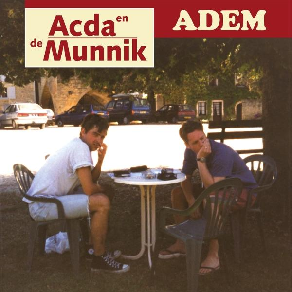 Zwei Männer sitzen im Freien an einem Tisch. Autos und ein Gebäude sind im Hintergrund. Der Text "Acda en De Munnik" steht oben.