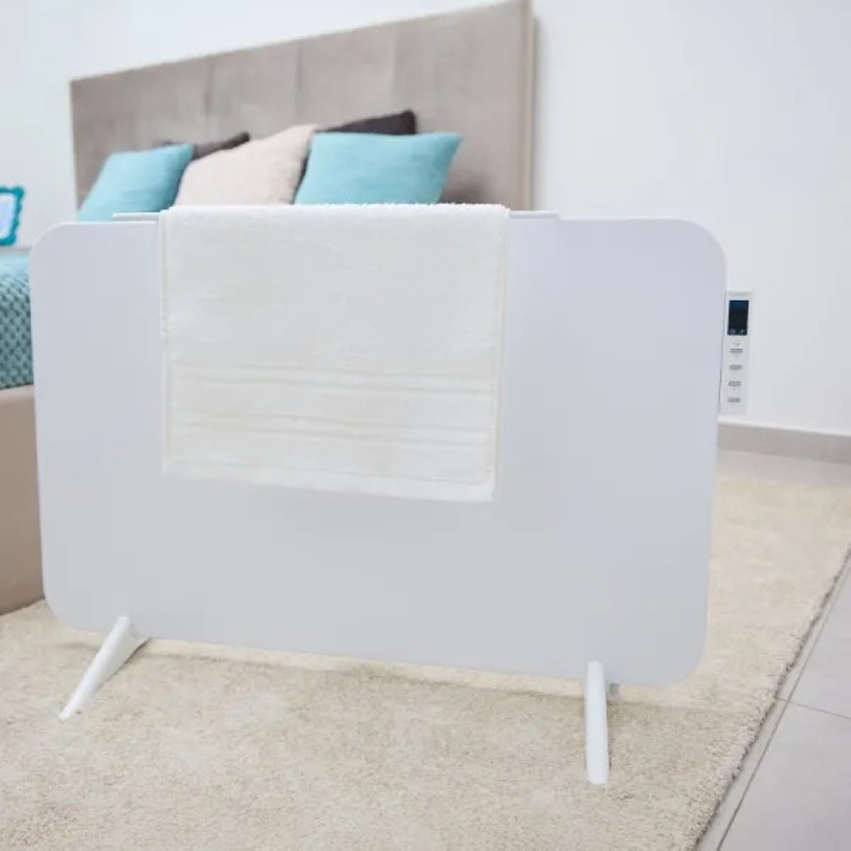 Biały ręcznik wisi na białym grzejniku. W tle łóżko z poduszkami. Podłoga pokryta jest beżowym dywanem.