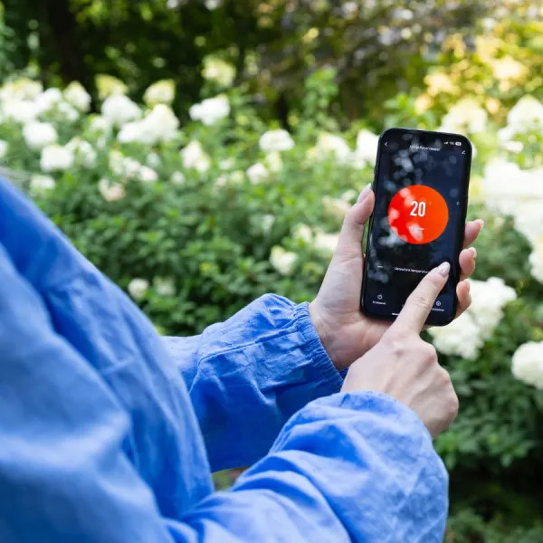Osoba trzyma telefon pokazujący 20 stopni. Osoba jest w ogrodzie z białymi kwiatami i zielonymi liśćmi.