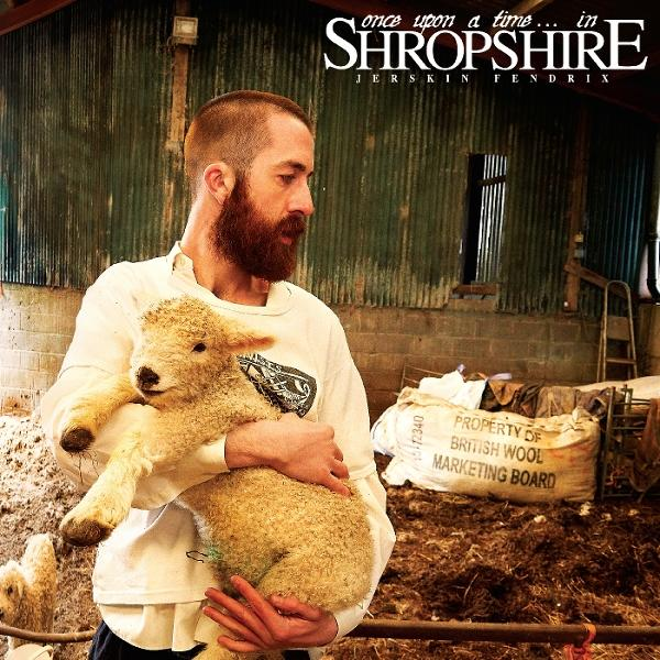 Ein bärtiger Mann hält ein Lamm in einer Scheune. Ein Schild zeigt 'British Wool Marketing Board'. Im Hintergrund sind eine Leiter und Heu zu sehen.