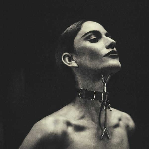 Eine Frau mit Lederhalsband und Halsfessel blickt nach oben. Dunkler Hintergrund. Schwarzer Lippenstift, kurze Haare und sichtbare Schultern.