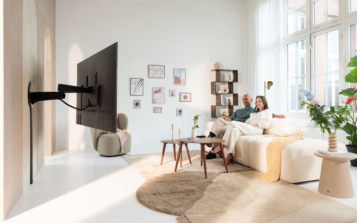 Salon z parą na kanapie, telewizorem na ścianie, sztuką i dekoracjami. Zawiera dywan, stoliki i rośliny. Jasny, nowoczesny design.