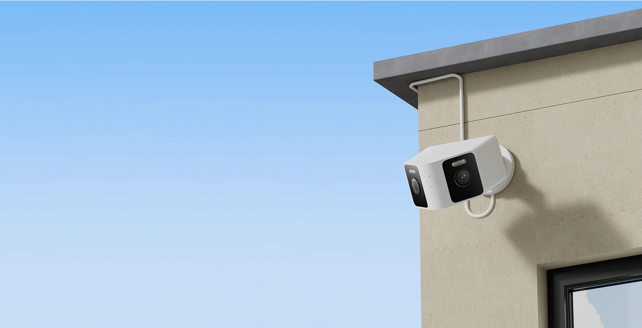 Kamera bezpieczeństwa jest zamontowana na zewnątrz budynku. Kamera jest biała z czarnym obiektywem. Niebo jest niebieskie.