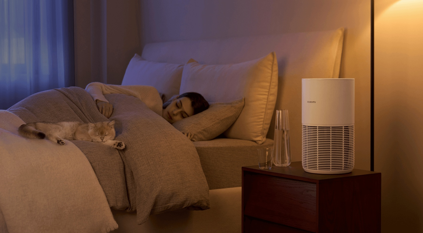 Kobieta i kot śpią na łóżku. Oczyszczacz powietrza stoi na stoliku nocnym, obok szklanki i karafki z wodą.