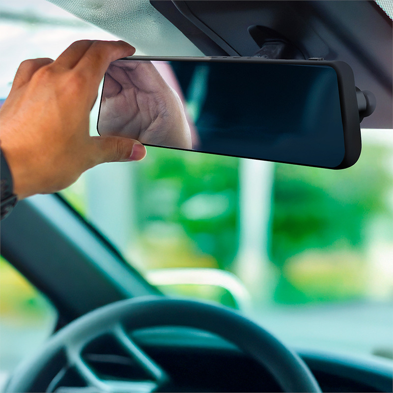 Ręka reguluje lusterko wsteczne samochodu. Odbicie pokazuje dłoń. Kierownica widoczna na pierwszym planie. Zielone drzewa w tle.