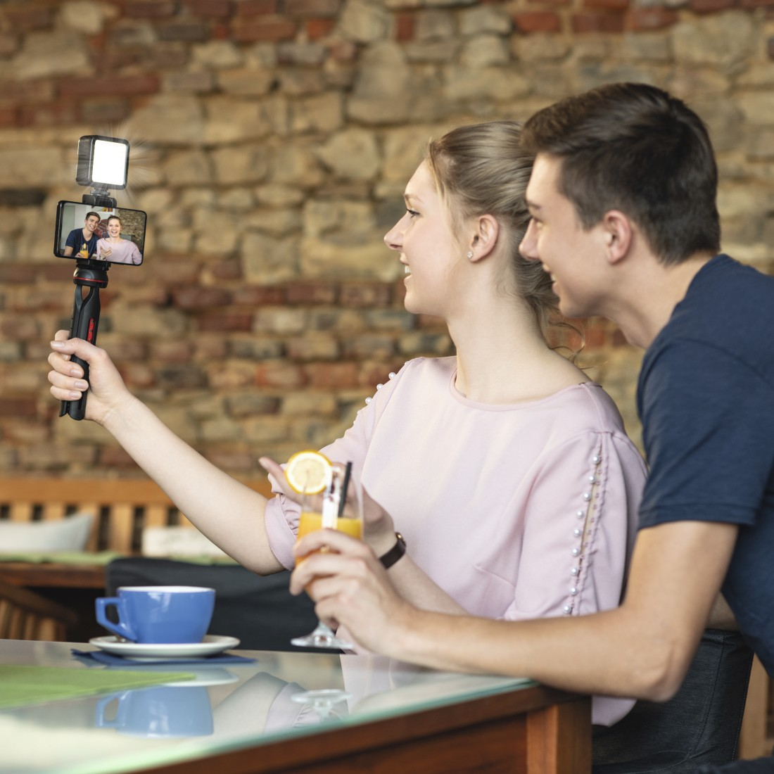 Bir çift kafede selfie çekiyor. Kadın, bir tripod üzerinde telefon tutuyor. Masada içecekler ve bir kahve fincanı var.