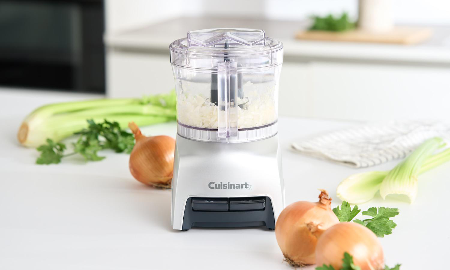Ein Cuisinart-Foodprozessor mit gehacktem Gemüse. Zwiebeln, Sellerie und Petersilie sind auf einer weißen Oberfläche angeordnet.