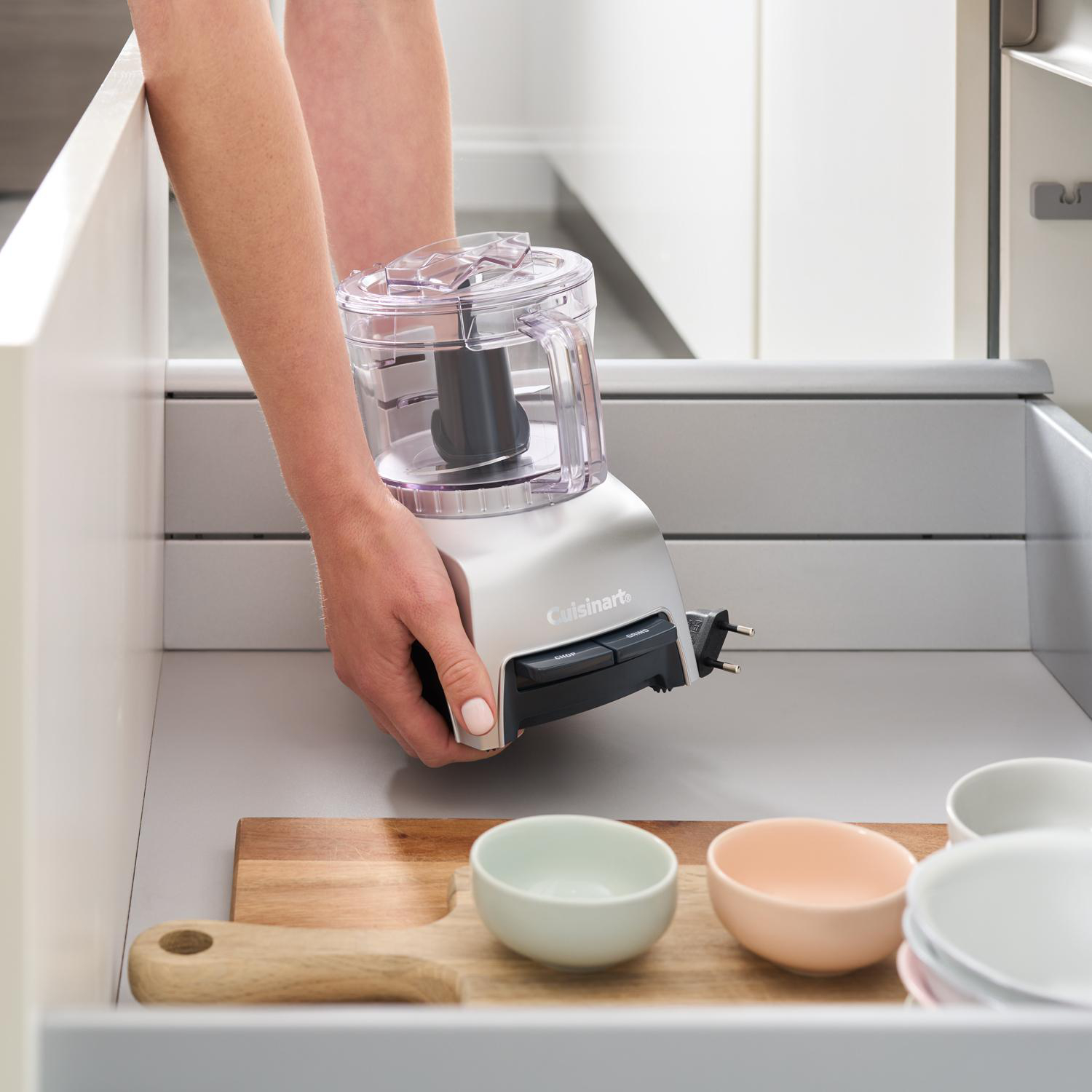 Eine Person legt einen Cuisinart-Foodprozessor in eine Schublade. Schüsseln und ein Schneidebrett befinden sich in der Schublade. Im Hintergrund sind weiße Schränke zu sehen.