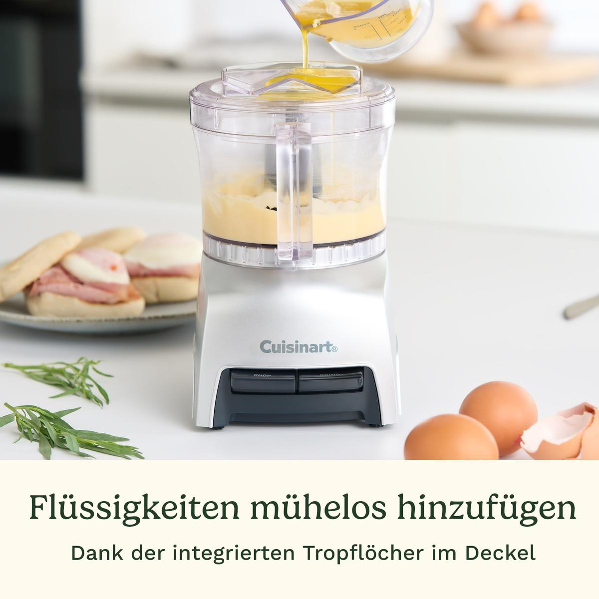 Ein Küchengerät, in das Flüssigkeit gegossen wird. Eier, Sandwiches und Kräuter liegen auf dem Tisch.