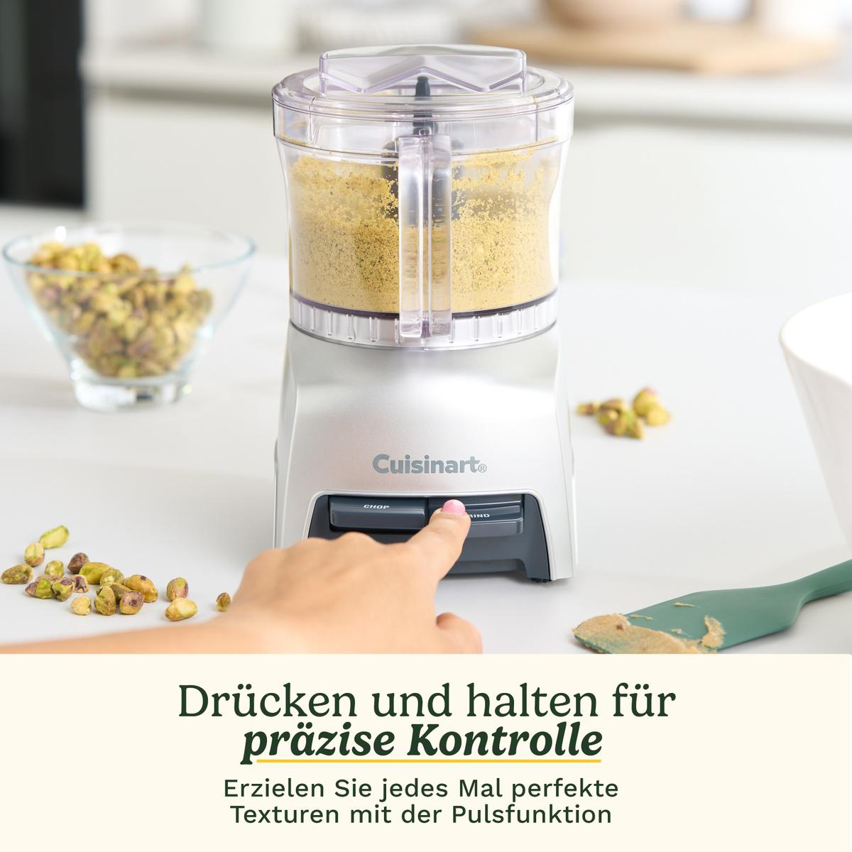 Eine Hand drückt eine Taste an einem Cuisinart-Foodprozessor. Pistazien und eine Schüssel stehen auf dem Tisch. Text sagt 'Präzise Kontrolle'.