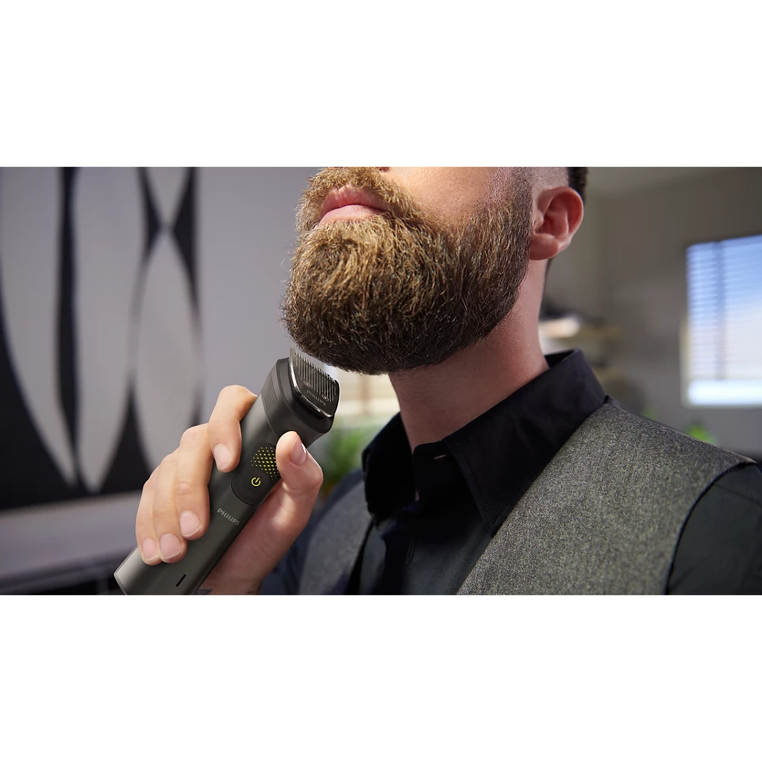 Un uomo taglia la barba con un regolabarba Philips. Indossa una camicia nera e un gilet grigio. Lo sfondo ha un muro a motivi e una finestra.