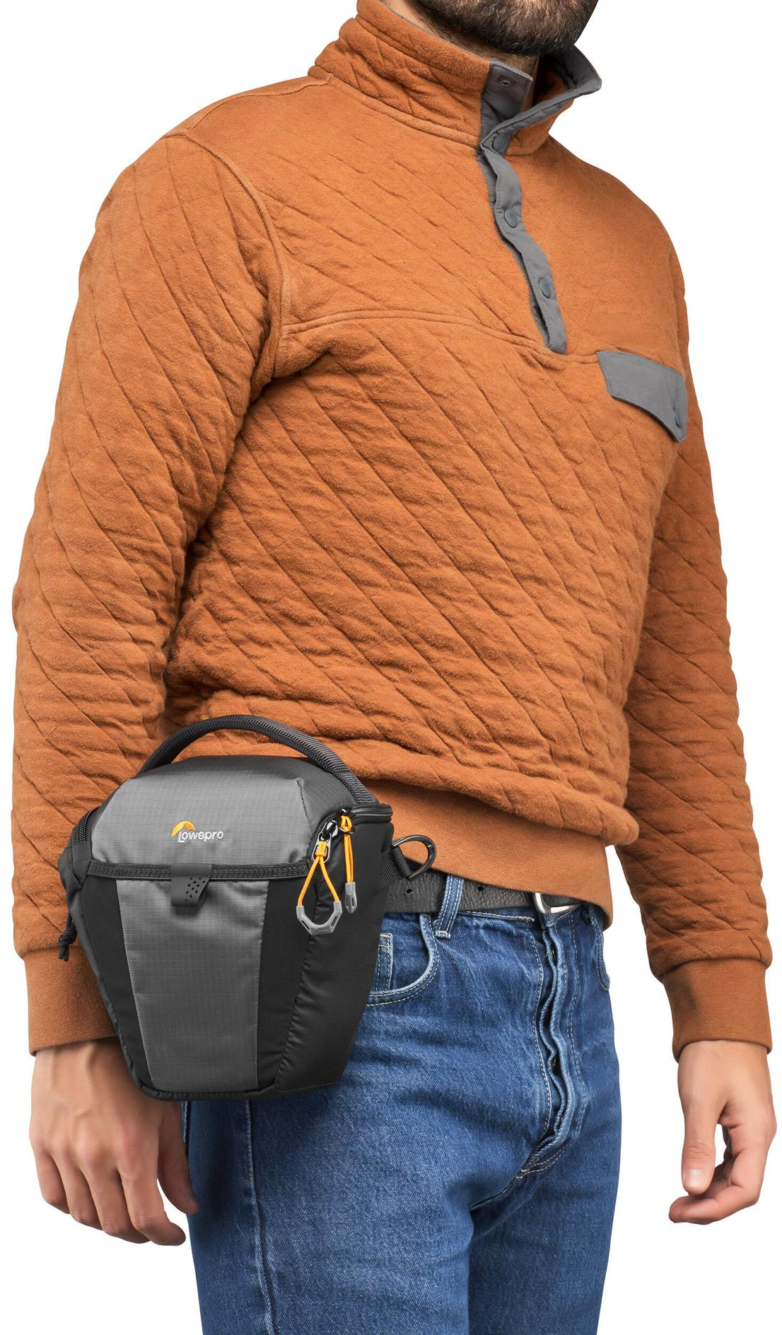 Un uomo con un maglione arancione e jeans tiene in mano una borsa per fotocamera Lowepro grigia e nera. La borsa è attaccata alla cintura.