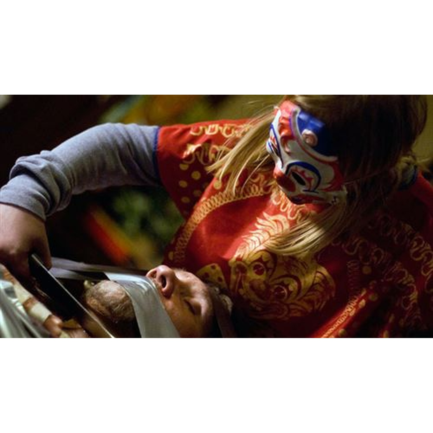 Une personne portant un masque et une robe rouge tient un outil métallique près d'une personne allongée. La personne est couverte d'un tissu blanc.