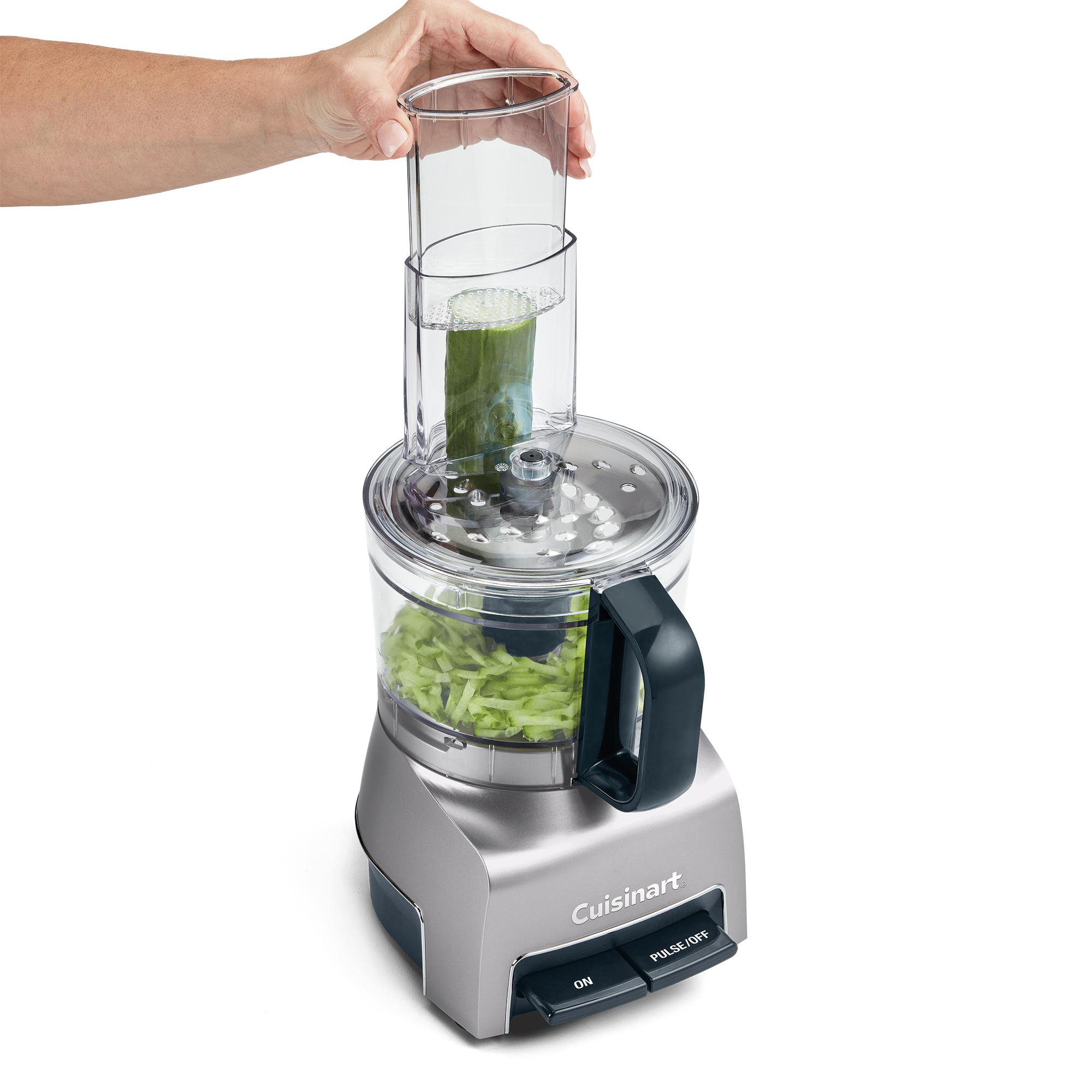 Eine Hand hält einen Behälter mit einer Gurke über einer Cuisinart-Küchenmaschine mit geschnittener Gurke.