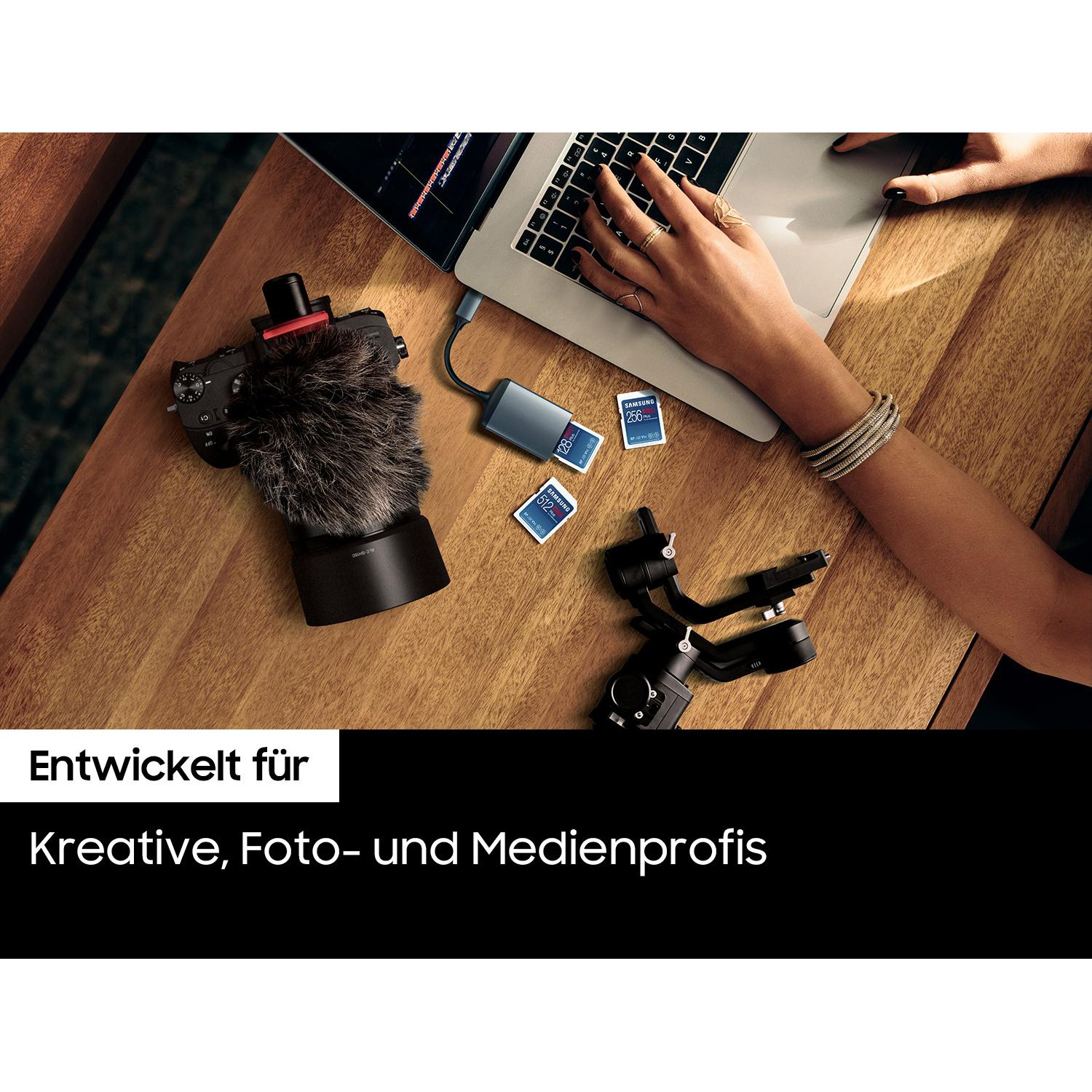 A wooden table with a camera, microphone, laptop, SD cards, and a person's hand with bracelets and rings.