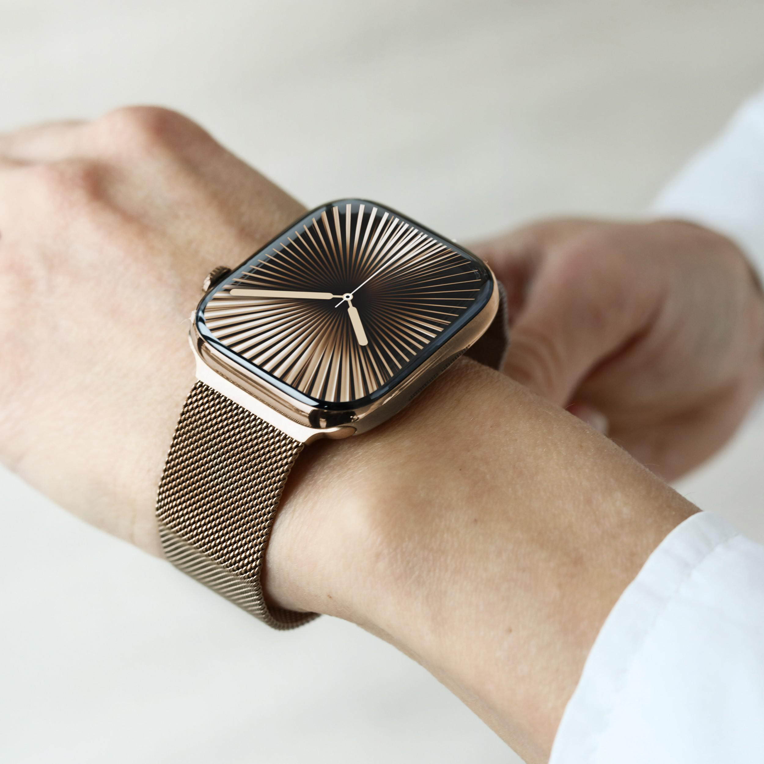 Eine Person, die eine goldene Uhr mit einem gemusterten Zifferblatt und einem goldenen Mesh-Armband am Handgelenk trägt, weißes Hemd im Hintergrund.