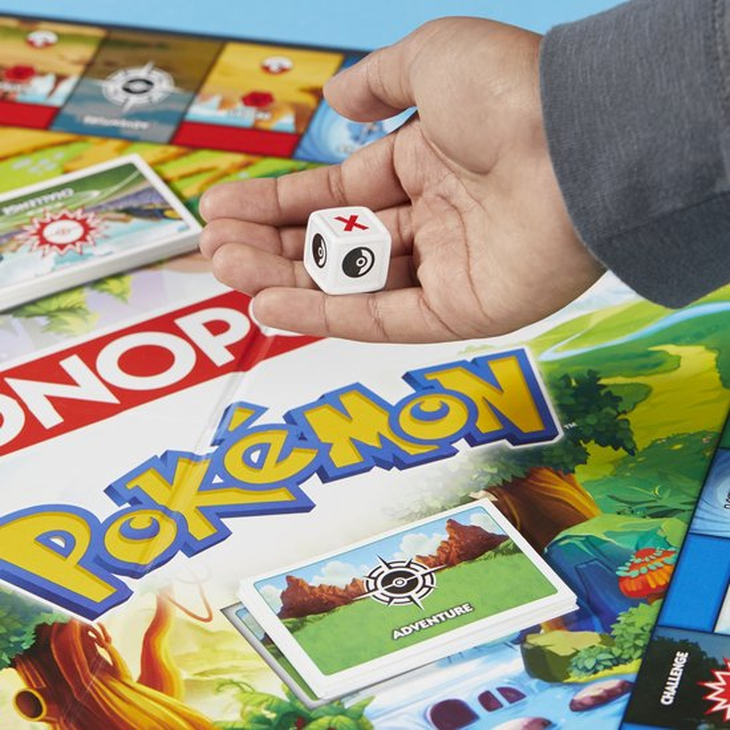 Hand die een witte dobbelsteen met zwarte symbolen vasthoudt, boven een Pokémon Monopoly bord. Kaarten en bord zichtbaar.