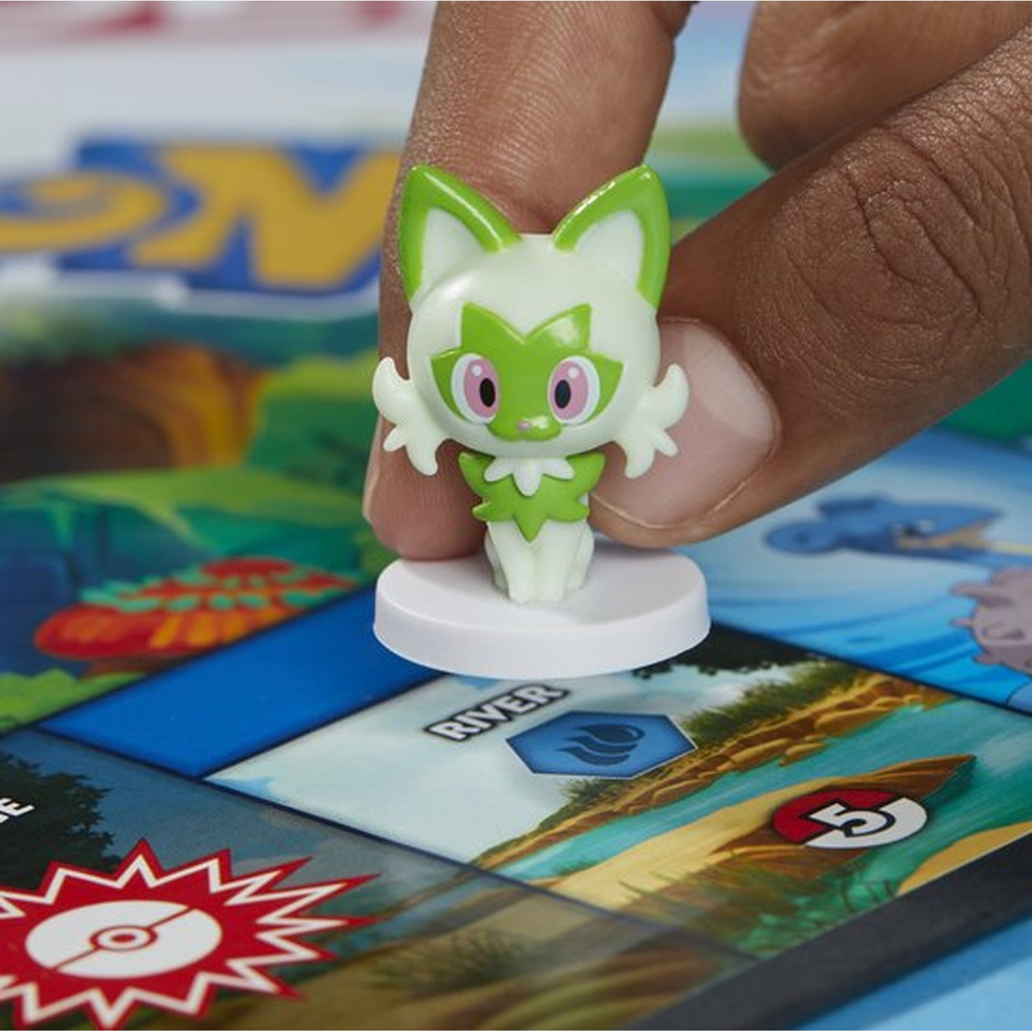 Een hand houdt een groen en wit Sprigatito-figuur op een witte basis. Het bevindt zich boven een kleurrijk bordspel met verschillende ontwerpen.