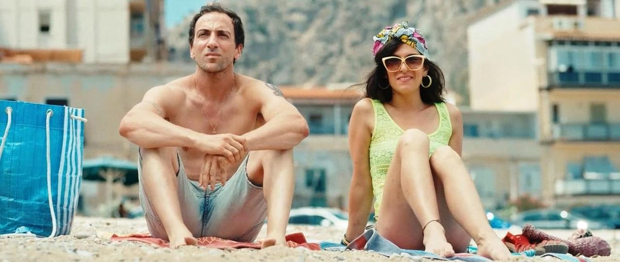 Ein Mann und eine Frau sitzen am Strand. Der Mann ist oben ohne. Die Frau trägt ein gelbes Oberteil, Sonnenbrille und ein Kopftuch. Gebäude im Hintergrund.