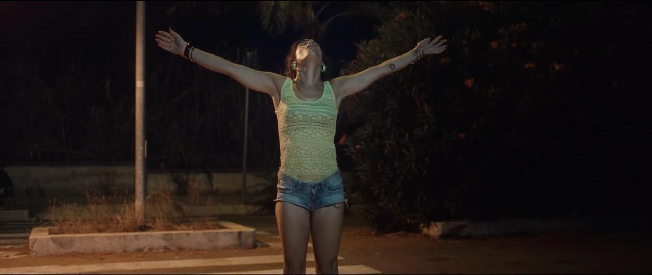 Eine Frau mit ausgestreckten Armen steht nachts auf einer Straße. Sie trägt ein gelbes Oberteil und Jeansshorts. Dunkles Laub umgibt sie.