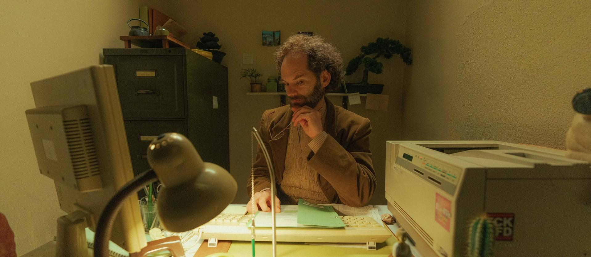 Ein Mann mit lockigem Haar sitzt an einem Schreibtisch. Er betrachtet Dokumente und benutzt einen Computer. Es gibt eine Lampe und einen Drucker.
