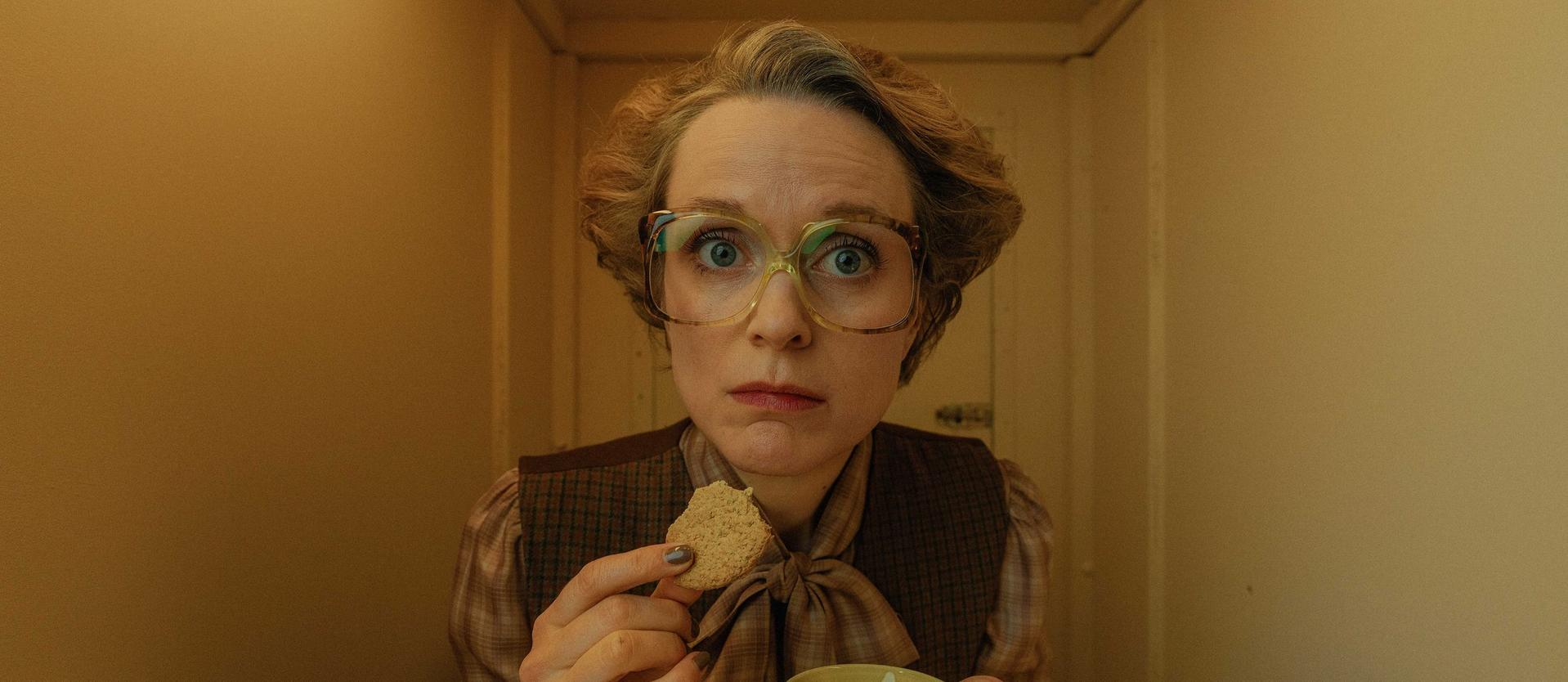 Eine Frau mit Brille und besorgtem Gesichtsausdruck hält einen Keks. Sie trägt eine braune Weste. Der Hintergrund ist beige.