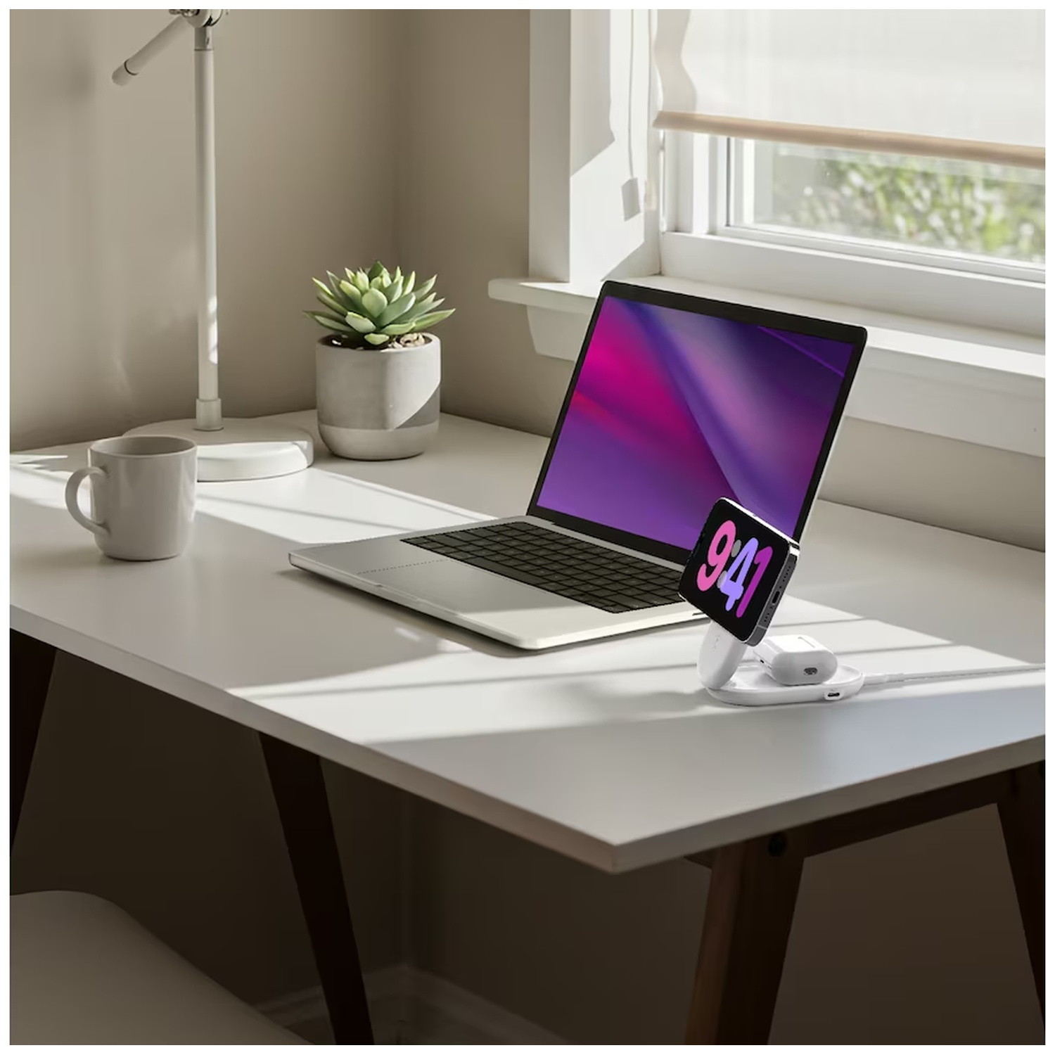 Un ordinateur portable, une montre et des AirPods sur un bureau blanc. Une plante succulente en pot et une lampe sont également présentes. Une fenêtre est en arrière-plan.