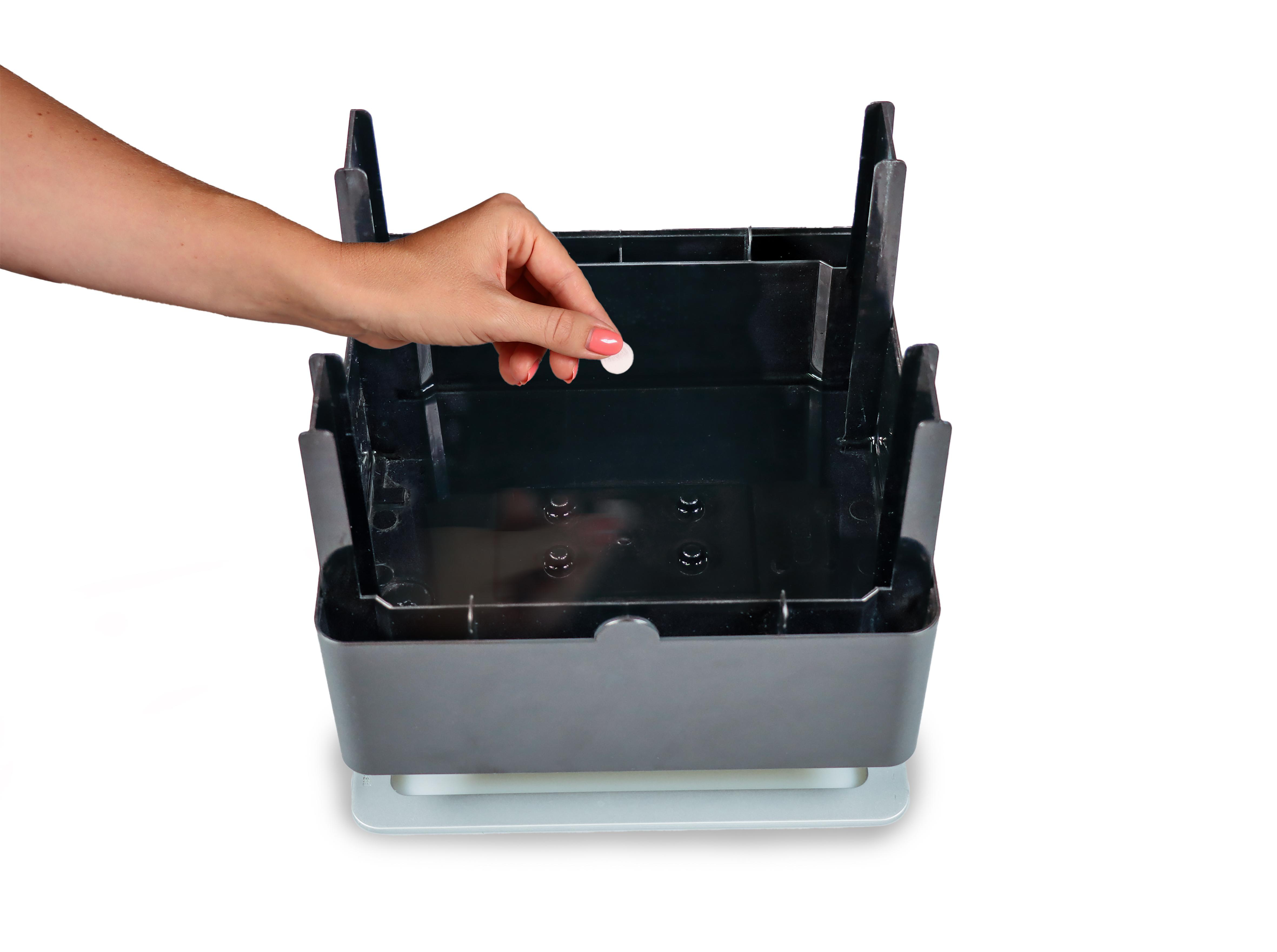 Eine Hand lässt eine kleine weiße Tablette in einen schwarzen Behälter fallen, auf weißem Untergrund.