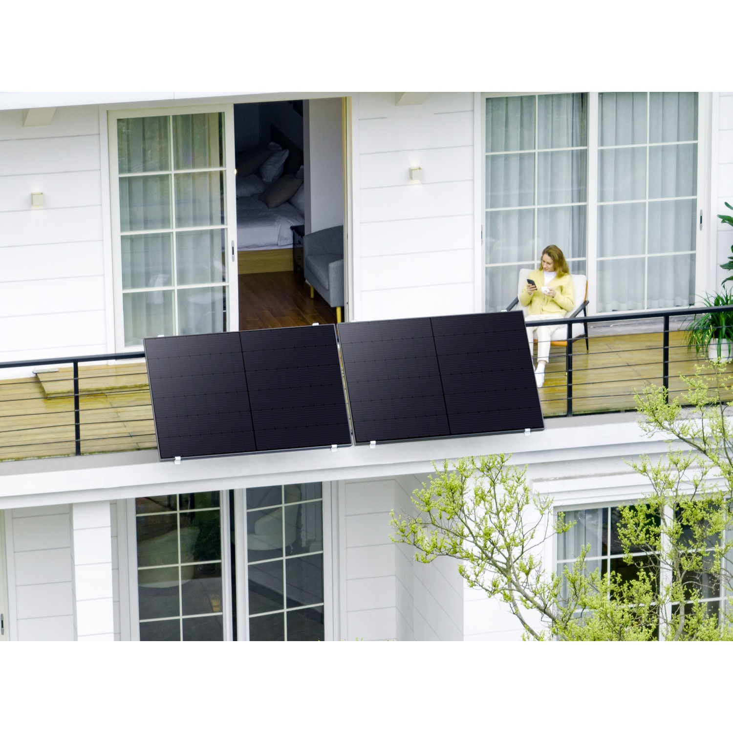 Une femme sur un balcon avec des panneaux solaires, utilisant son téléphone. Le balcon fait partie d'une maison blanche, des arbres au premier plan.