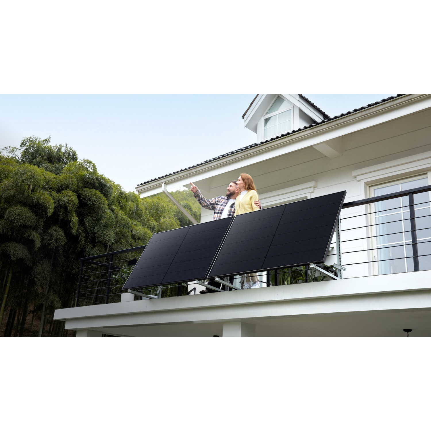 Un couple sur un balcon pointe vers le ciel. Des panneaux solaires sont installés sur le balcon. Des arbres verts entourent la maison.