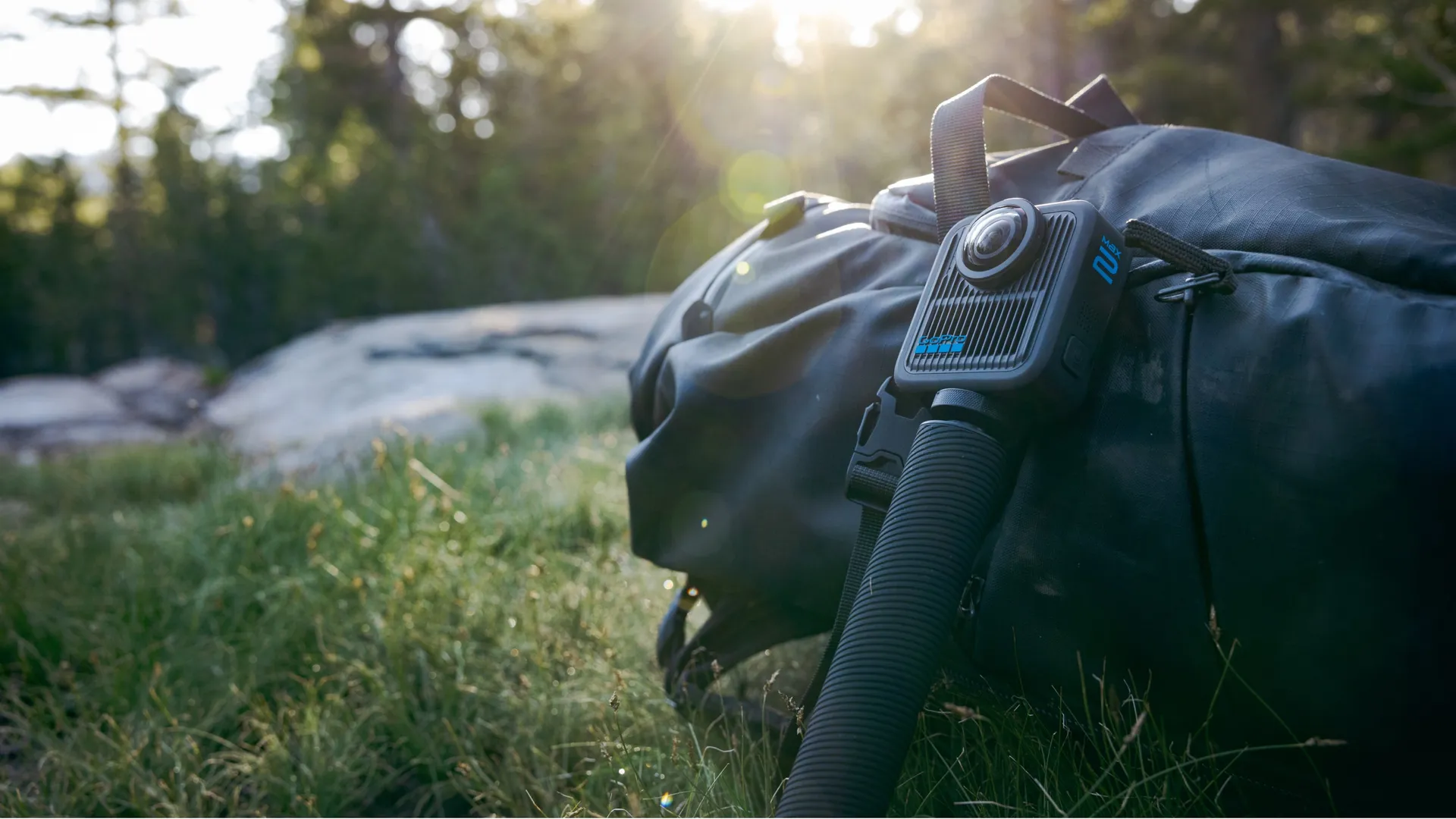 Egy GoPro Max 360 kamera egy fekete hátizsákhoz van rögzítve. A háttérben fű és erdő látható.
