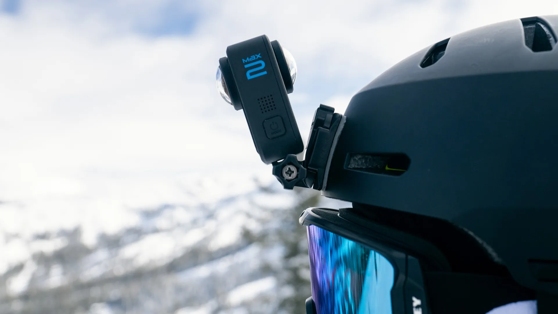 Egy fekete GoPro Max 2 kamera egy fekete sisakra van szerelve. Hóborította hegyek és felhős ég a háttérben.