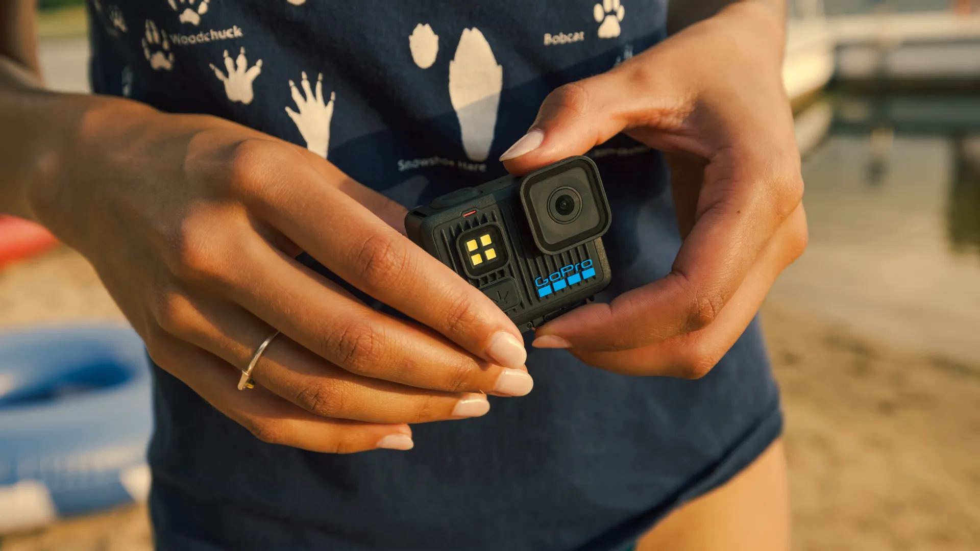 Egy személy egy GoPro kamerát tart. A kamera fekete, kék felirattal. A személy kék inget és gyűrűt visel. A háttér elmosódott.