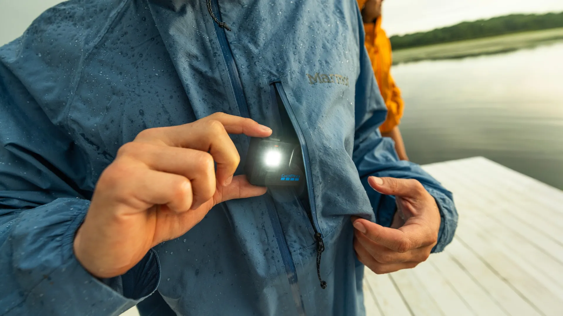 Kék kabátos személy GoPro kamerát tart, ami fényesen ragyog. Vízcseppek a kabáton. Háttérben víz és stég látható.
